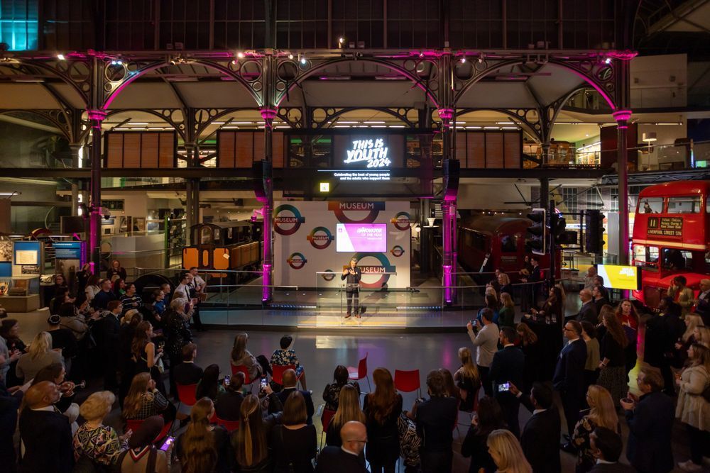 This is Youth event at the London Transport Museum featuring a speaker addressing an audience with red double-decker buses and a London Underground sign in the background.