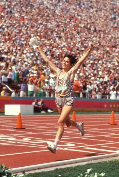 photo of Joan Benoit as she crosses the line at the Olympics. She's a white woman with dark hair and her arms aloft.