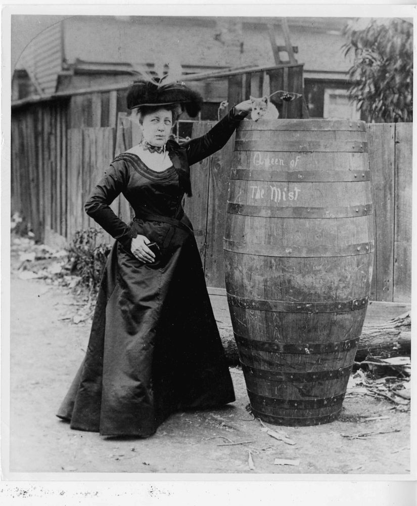 Annie Edson Taylor standing with the barrel "Queen of the Mist". She is a white woman in a floor-length skirt, dark jacket and a big hat. A small pale cat is sitting on top of the barrel.