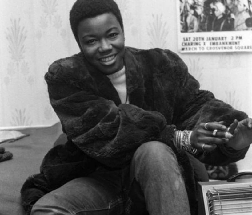 Olive Morris in her squat in the 1970s. She is a black woman with a short fro, wearing a big fluffy coat as she tries to keep warm with a one bar fire. Photo by Neil Kenlock.