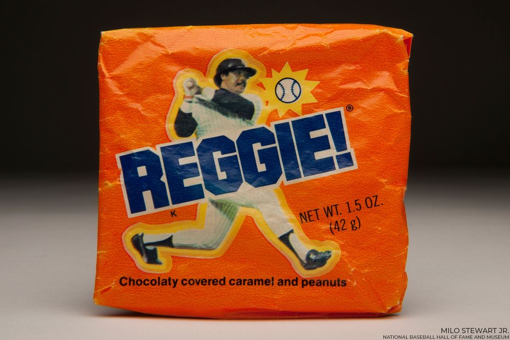 Blue REGGIE! text on an orange candy wrapper depicting Reggie Jackson midway through his left-handed swing. (Milo Stewart Jr./National Baseball Hall of Fame and Museum)