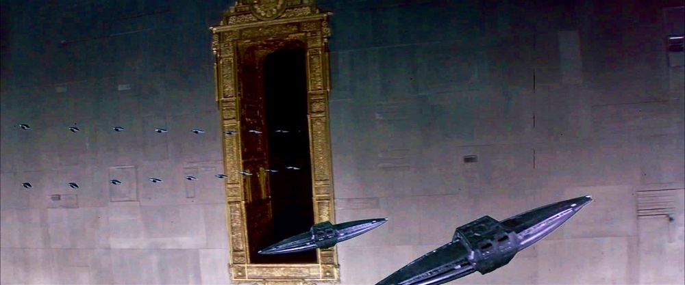 Oblong black Atreides spaceships enter the rectangular loading bay of a giant gray spaceship. The loading bay is ornately edged with a baroque gold frame. 