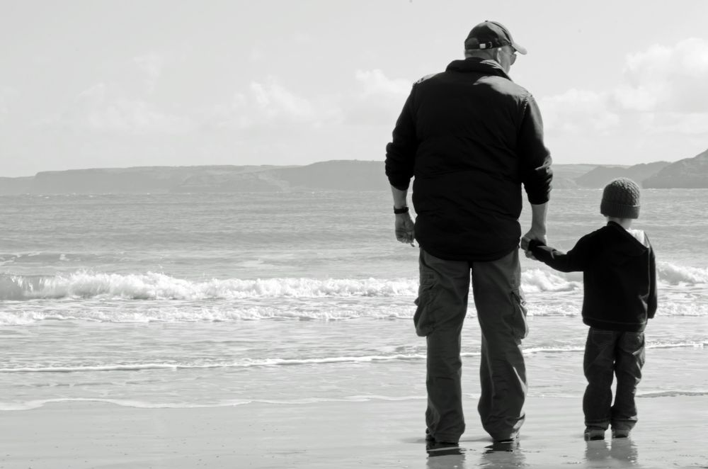 Black and white photo of a grandfather and his grandchild standing hand in hand on a beach, facing the ocean with waves gently rolling in. The adult is wearing a cap, a jacket, and cargo pants, while the child wears a knit hat, a hoodie, and jeans. In the background, there are distant hills under a partly cloudy sky.