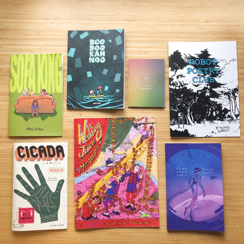 Seven brightly colored self-published comics displayed on a bamboo table. Each comic is a different size and the lush drawings that adorn them indicate their depth and breadth of styles comics can come in!