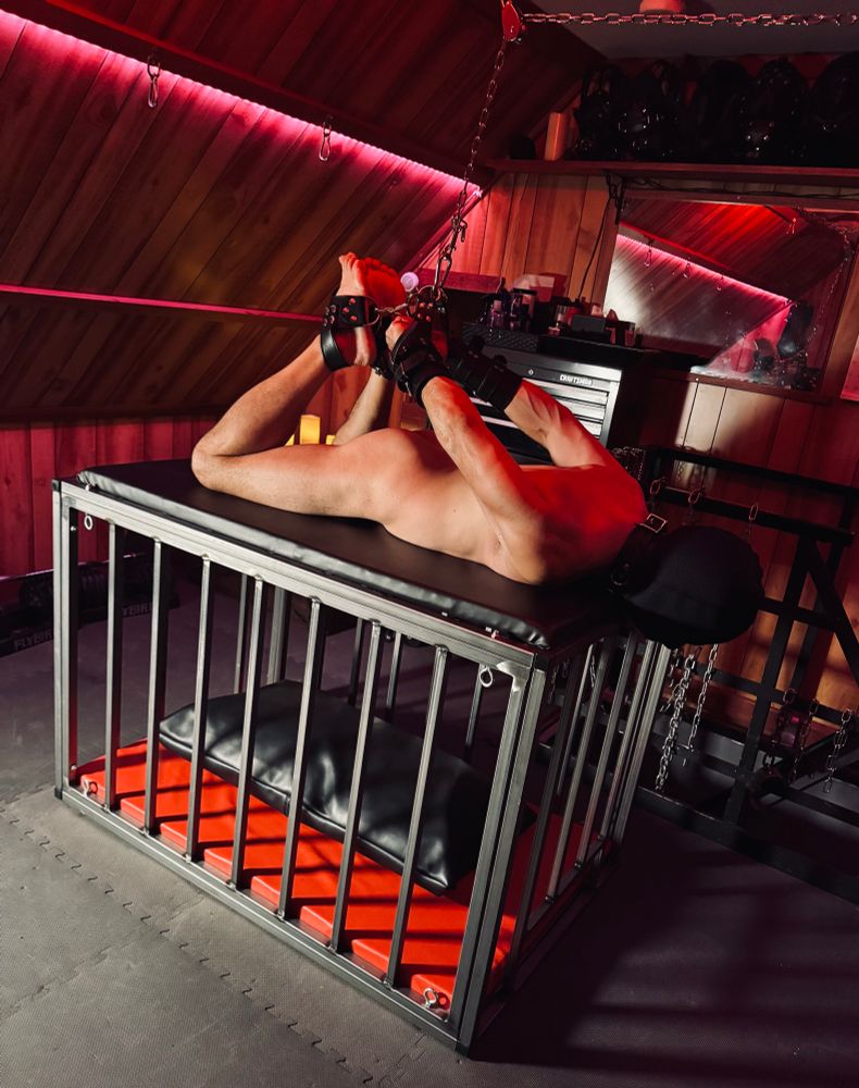 Naked male sub laying across a metal cage in a dungeon, hogtied with legs and arms suspended by a chain