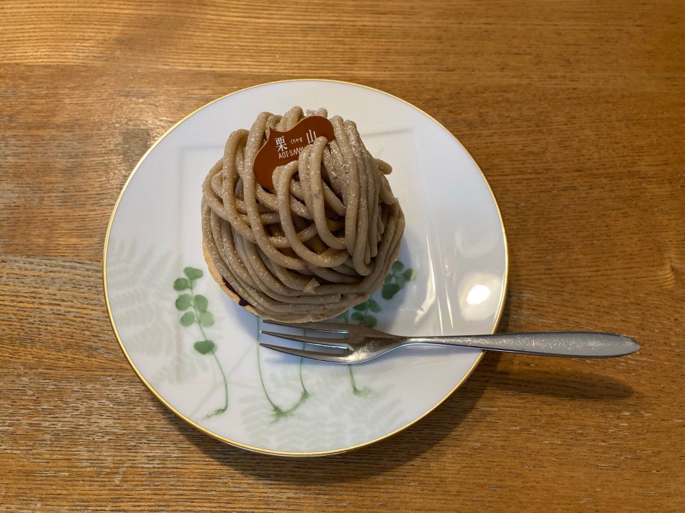 丸い小皿(白地に金縁、植物の絵が描かれている)にのせられたモンブランと銀色のフォーク。モンブランの頂上には「栗山」と書いた紙が刺さっている。