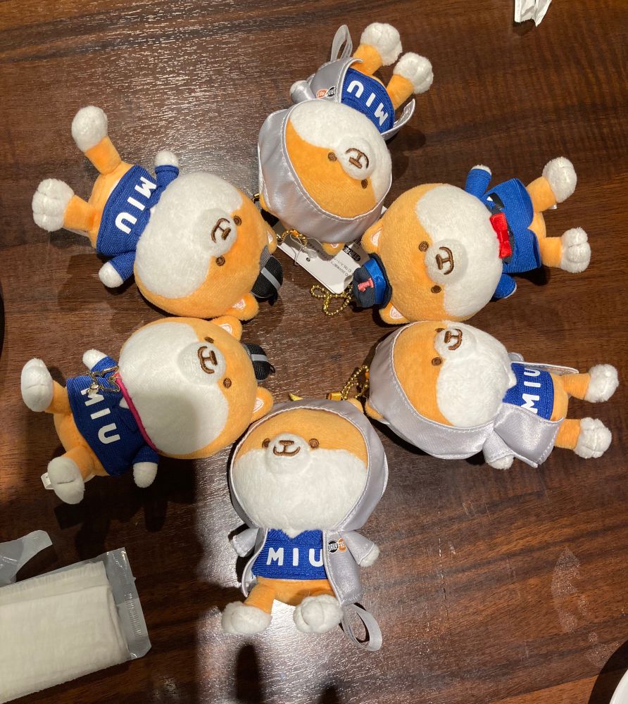 テーブルの上に並べられたポリまるキーチェーンぬいぐるみ6つ。頭を中心に、足を外側に向けて円を描いている。
ポリまるは、ドラマ「MIU404」のオレンジと白色の犬のキャラクター。