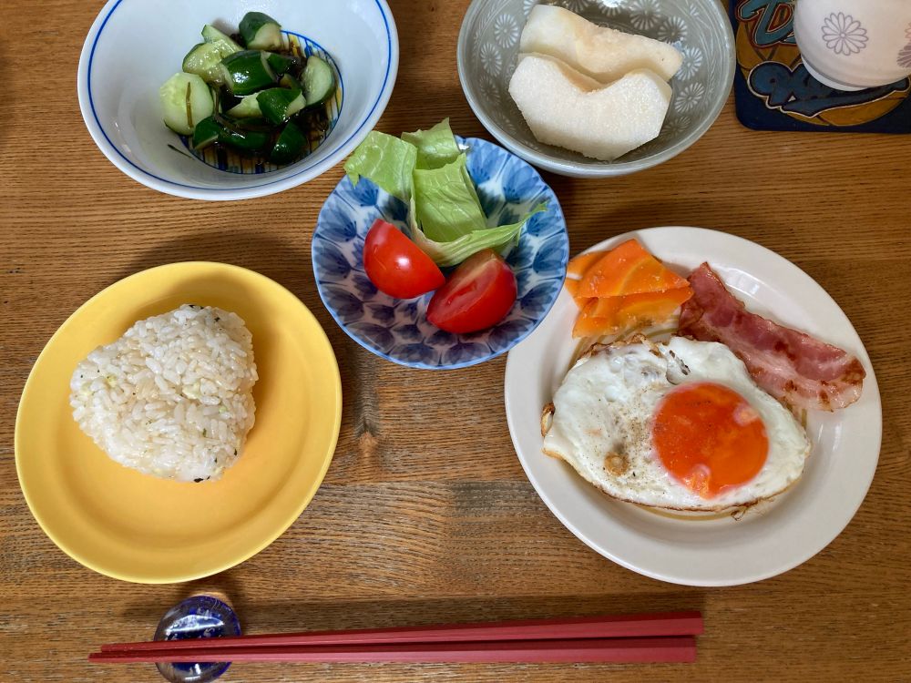 食卓の写真。小皿や小鉢に、おにぎり・目玉焼き・トマトとレタス・きゅうりの漬物・梨がのっている。