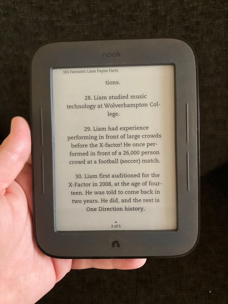 My hand holding up an old Nook ereader with a Liam Payne fan book on the screen