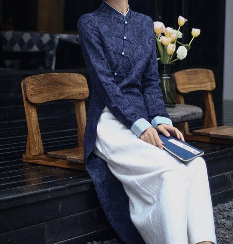 A woman wearing a dark blue qipao with white pants underneath. She is sitting and holding a book.
