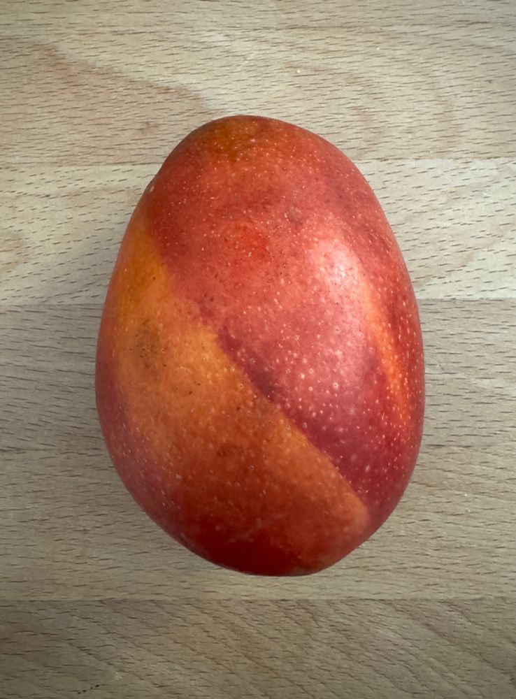 A round mango with a very vibrant dark orange colour and swirl patterns on the skin, sitting on a plain wooden table