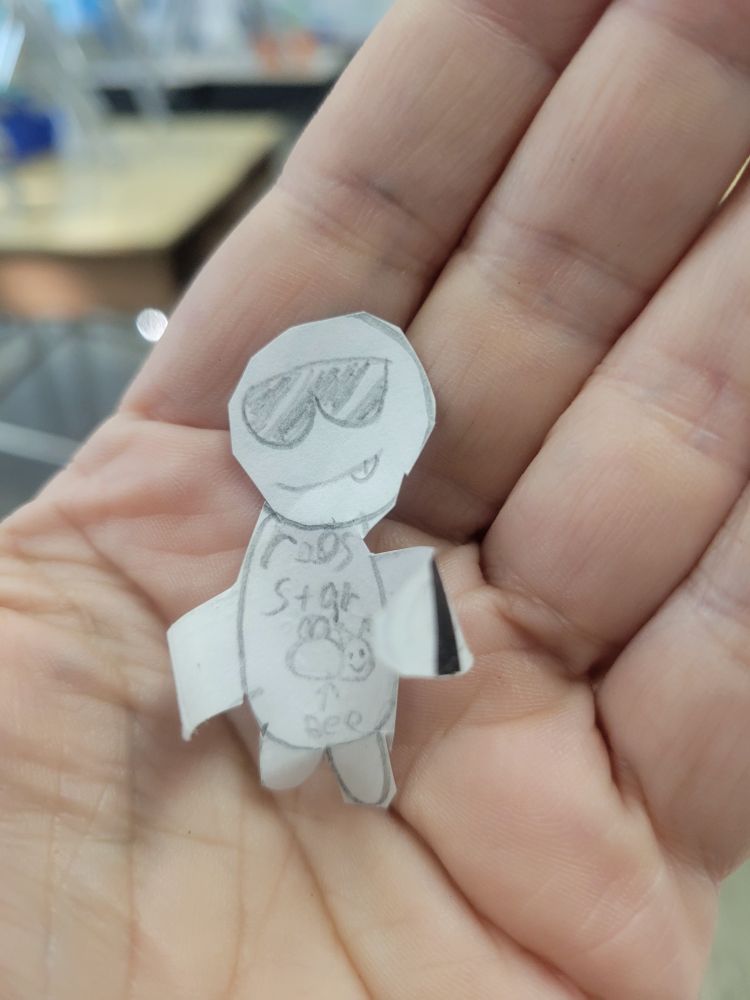 A small paper "Rhys Darby" drawn by a 4th grader wearing a shirt that states "roos star bee" with a picture of a smiling bee.  It's arms are curled so you can wear it on your pinky.