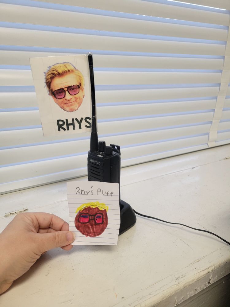 Photo of a black walkie talkie with a flag attached to the antenna that says 'RHYS' with a picture of Rhys Darby's head.  A hand is holding a small piece of paper with a child's drawing of the Rhys Darby photo with the words 'Rhy's Puff' written above the drawing.
