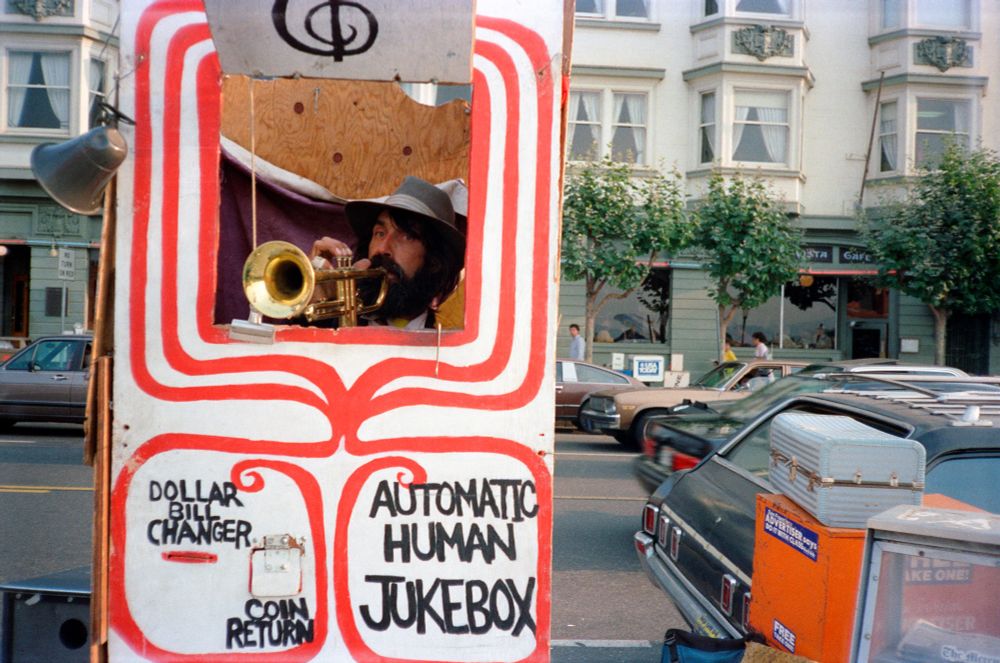A bearded street performer with a trumpet sits inside a big, painted refrigerator box, wearing a fedora. The box is mostly white, but has painted red ribbons around the window where he plays and a slot where you put in a dollar.