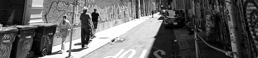 In a black and white photograph three people walk down a thin alley, into the harsh sunlight, casting shadows behind them. There is a lot of graffiti.