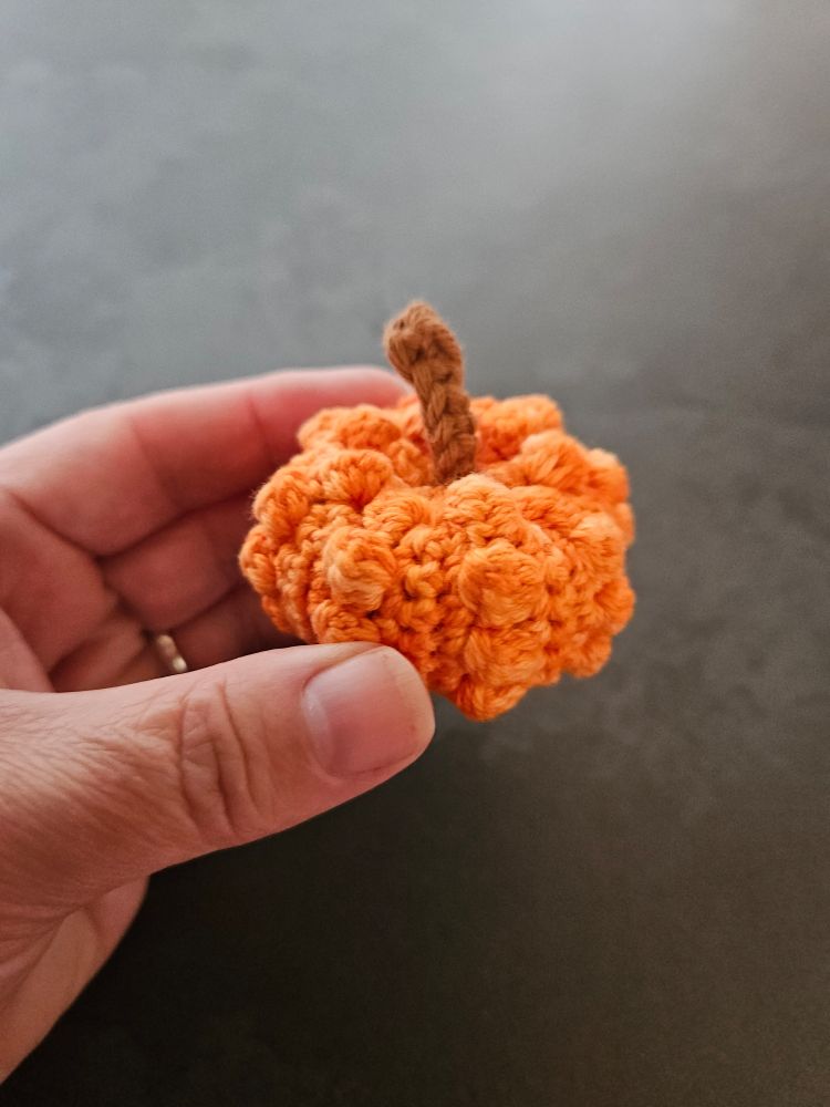 A little crocheted pumpkin held in a middle-aged white woman's hand to demonstrate that it's really quite small. It's knobbly and orange (the pumpkin, not the hand) with a brown stem.