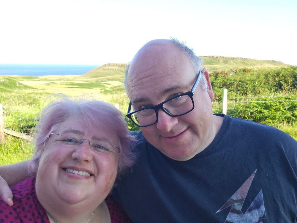 A selfie of Wendy and Greg. We're a middle-aged overweight white couple, both wearing glasses, and slightly sunburned on our noses and cheeks. Wendy has pink hair and Greg is mostly bald. We're standing in front of a lush green landscape, which is Treshnish on the west coast of the Island of Mull in the Scottish Hebrides. It's a beautiful sunny morning, and neither of us wants to leave.