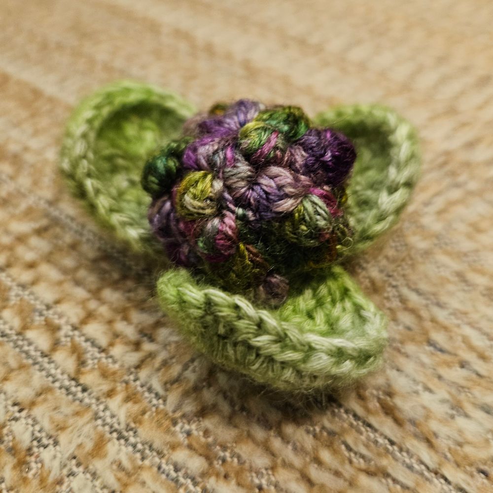 A photo of a tiny crocheted cauliflower. The head is made with a variegated yarn in shades of purple and green, and the leaves with a pale green variegated yarn. There's nothing in the picture to show scale, but the whole thing is about 2 inches across.