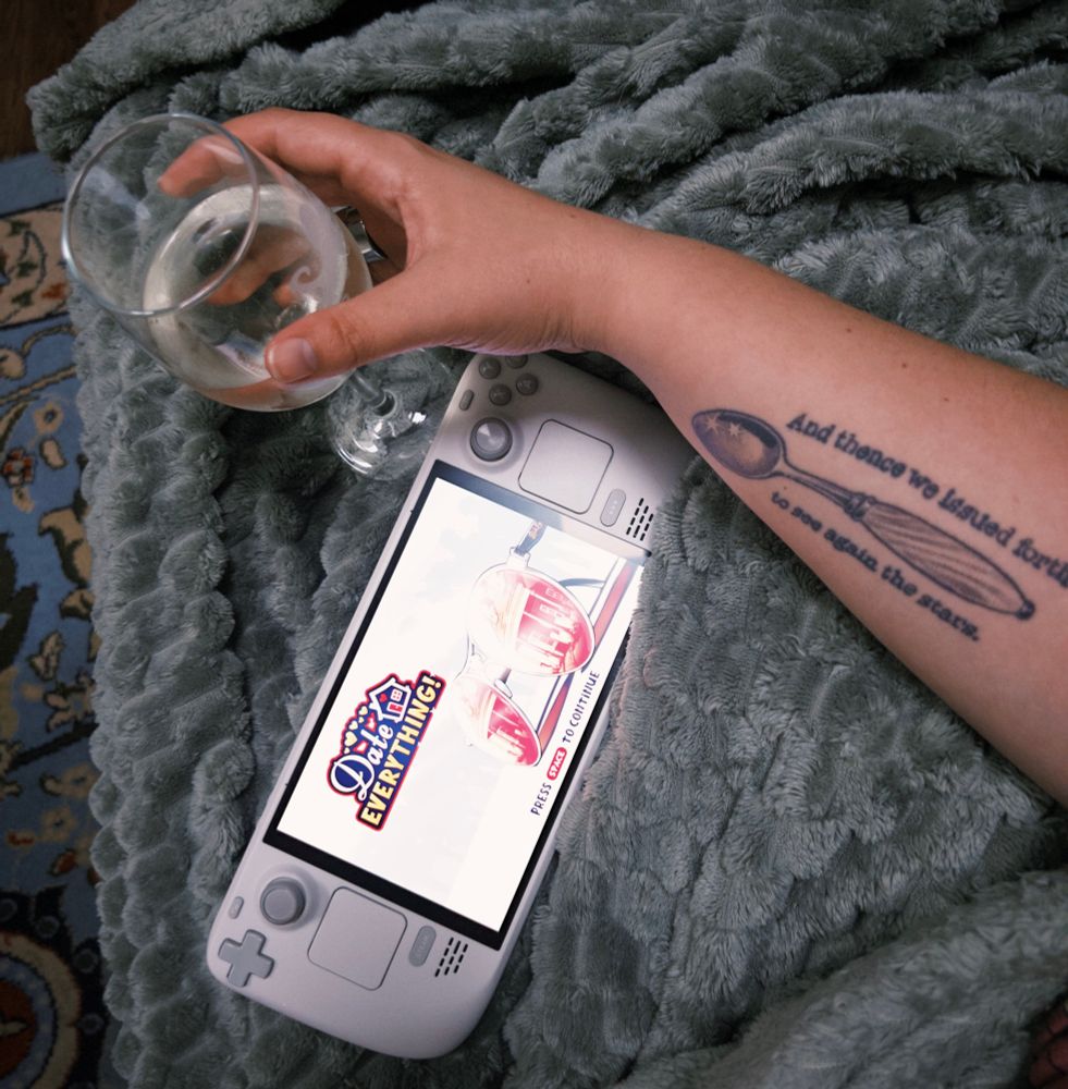 a top-down picture of a white steam deck with Date Everything open on screen. Ali’s right arm holds a glass of white wine in her hand. You can see a tattoo on the inside of her arm of a coffee spoon with word around it that read “And thence we issued forth to see again the stars”