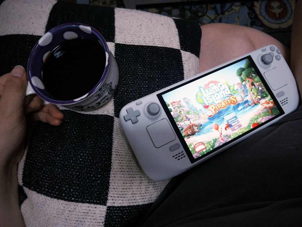 a top down picture of ali’s lap. on it rests a purple & white polka dot coffee cup full of black coffee, a black and white checkerboard pillow, and a white steam deck with luma island open on it 