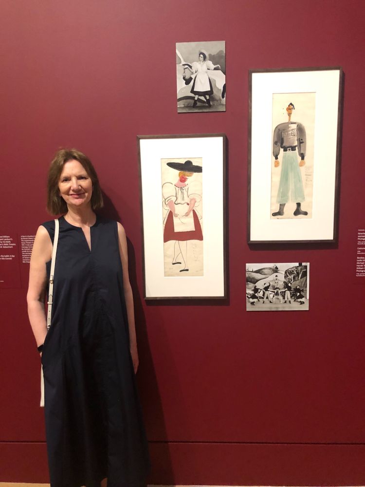 a woman in a blue dress stands in front of a maroon gallery wall, showing two costume paintings and two black and white photos of the costumes as worn by royal ballet dancers