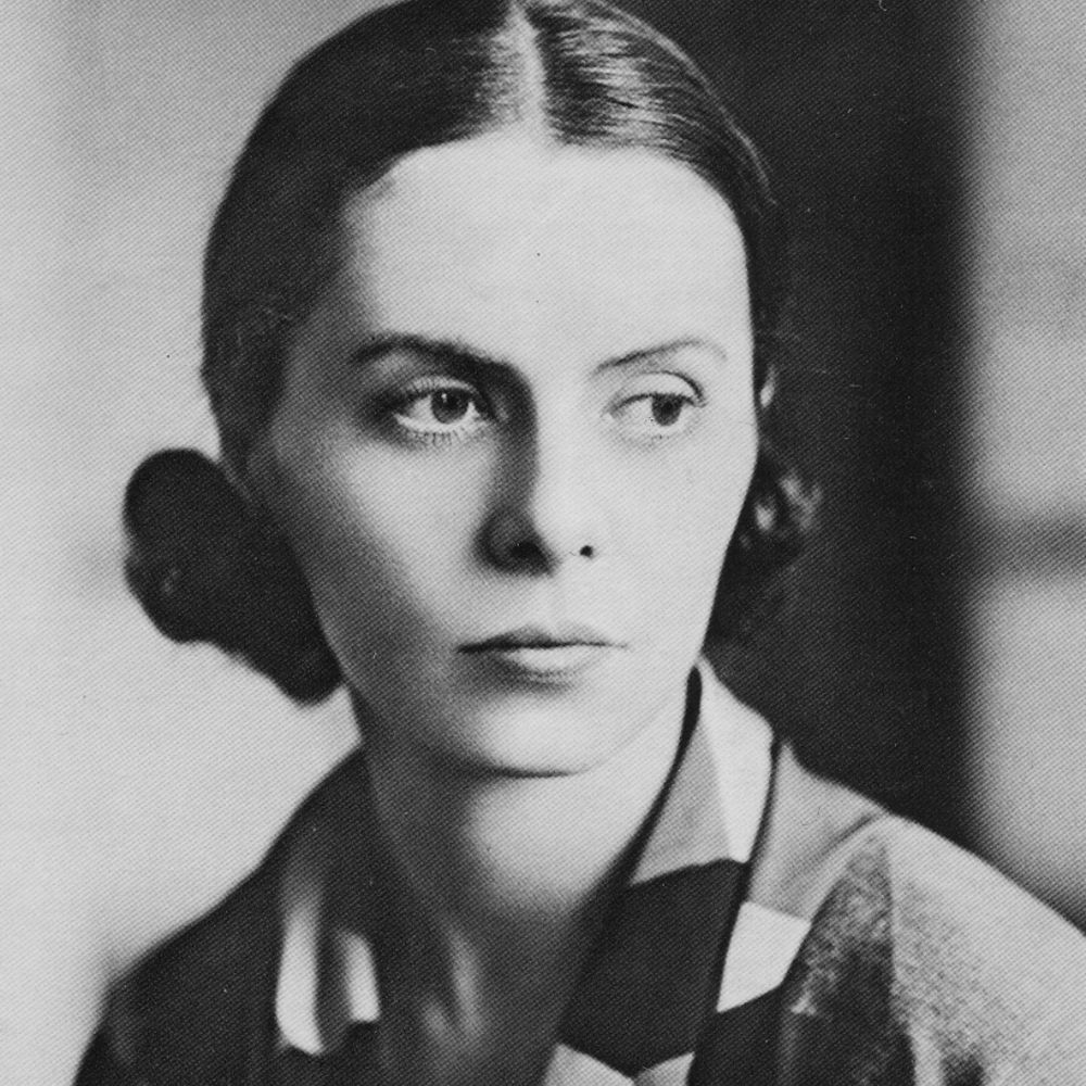 A vintage black and white portrait of a woman with dark, neatly parted hair pulled back into two low buns. She has a serious expression, high cheekbones, and a strong gaze directed slightly to the side. She is wearing a collared shirt with a patterned scarf draped around her neck. The background is blurred, and the lighting softly highlights her facial features.