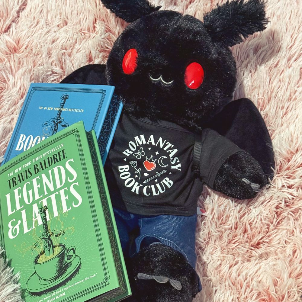 Square photo of a Build a Bear Mothman wearing a black t-shirt that says “Romantasy Book Club” and a pair of jeans. In front of the Mothman stuffie are 2 books: in the forefront a hardcover copy of Legends & Lattes by Travis Baldree and behind a hardcover copy of Bookshops & Bonedust, also by Travis Baldree.