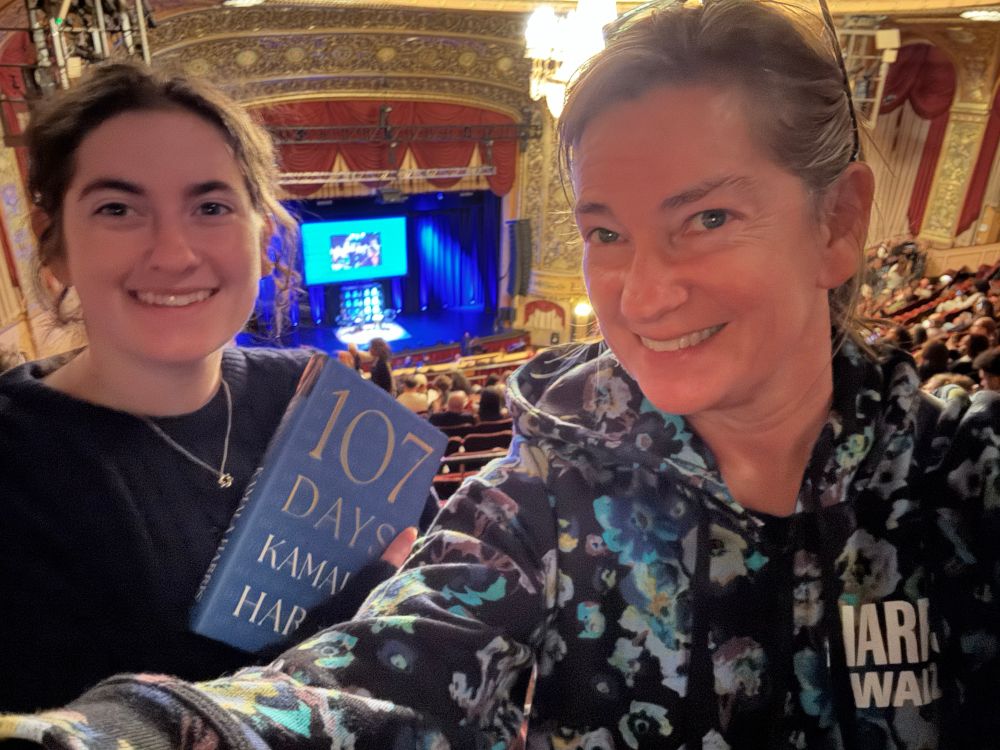 A young woman holding the Kamala Harris book with an older woman in a campaign hoodie in a balcony above a book talk stage. 