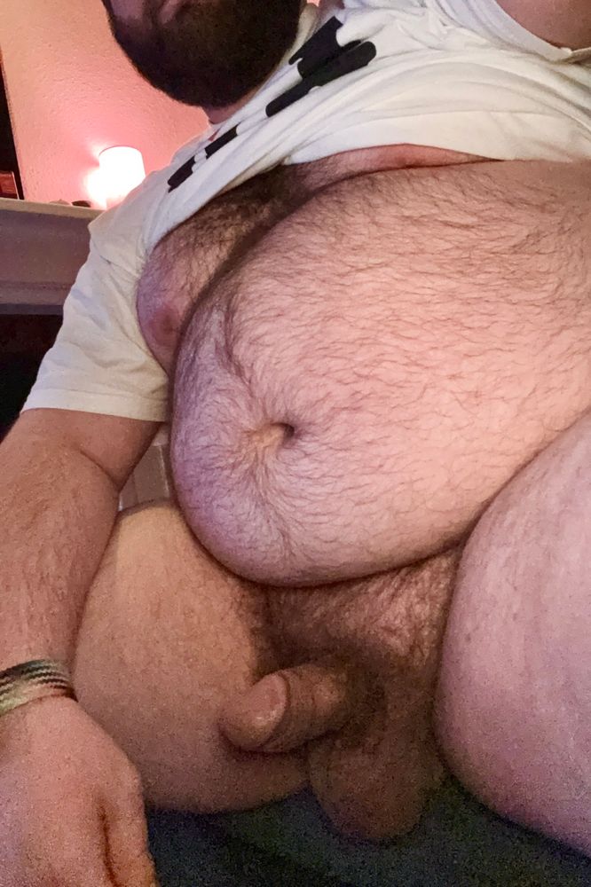 A fat, hairy bear has his shirt pulled up to show off his tummy and cock.