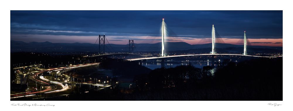 The Queensferry Crossing in the twilight with light trails s ing towards it. The main the Forth bridges can be seen the background. There are blue hues with an orange streak low on the horizon hinting at a sunset.