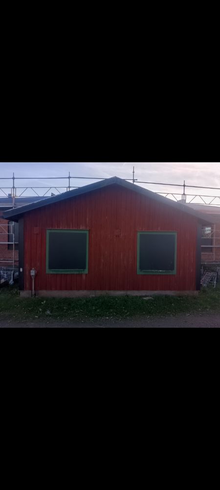 One side of a red wooden building with two windows which is covered up with black borads. Behind the red building is part of a brick buildind.