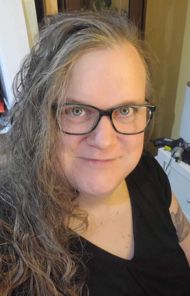 A nerdy white woman with very messy, wavy hair. She's looking at the camera and smiling.