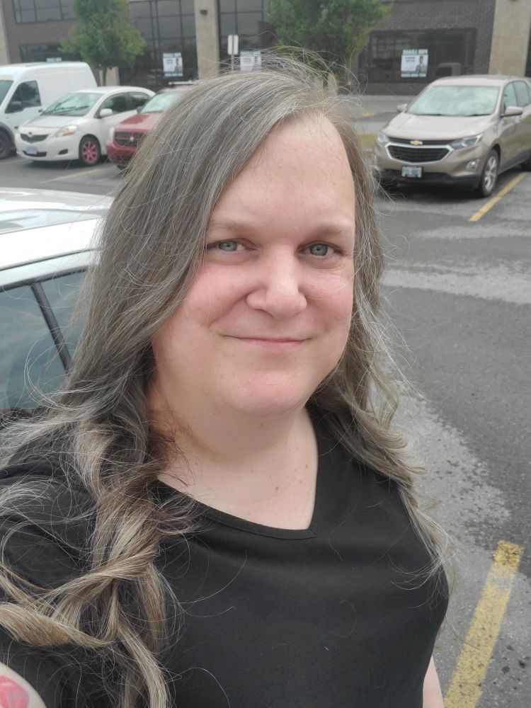 A white woman smiles at the camera while standing in a parking lot. Her beautiful brown and grey hair tumbles over her shoulder in loose curls