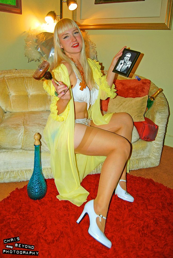 A photo of a blonde model wearing vintage lingerie including bra, panties, stockings, high heels and a yellow sheer robe. She is holding a picture of televangelist Pat Roberts in one hand and an almost empty glass of wine in the other. There is a blue vase near her on the floor and she is sitting on an old couch. There are lights and framed pictures behind her. There is a shag orange rug on the floor.