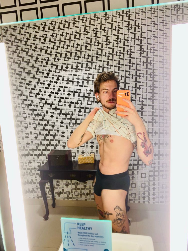 Gay guy with tattoos lifting his shirt and in a pair of black briefs 