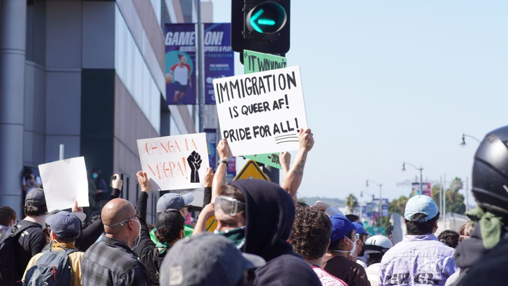 A crowd of protesters stands in the street under a green walk signal, holding signs in support of immigrant rights. One sign reads “IMMIGRATION IS QUEER AF! PRIDE FOR ALL!” and another says “CHINGA LA MIGRA!” with a raised fist symbol. People wear masks, bandanas, and caps, and a building in the background displays a “GAME ON!” banner.