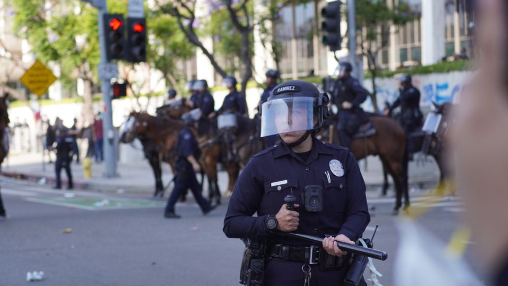 A line of LAPD officers in riot gear stands in formation in front of a building, with one officer prominently in the foreground holding a baton and wearing a face shield labeled “RAMIREZ.” Behind them, more officers on horseback and graffiti can be seen on the wall, along with scattered trash on the street.