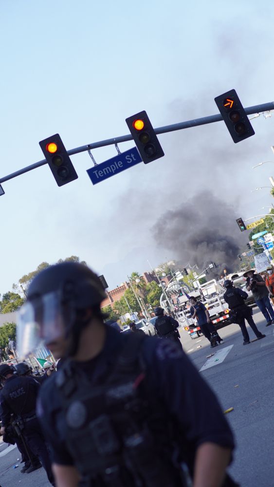 Police in riot gear are gathered near the intersection of Temple Street, where thick black smoke billows into the sky in the distance. Several officers are in the foreground. The scene appears tense and chaotic.