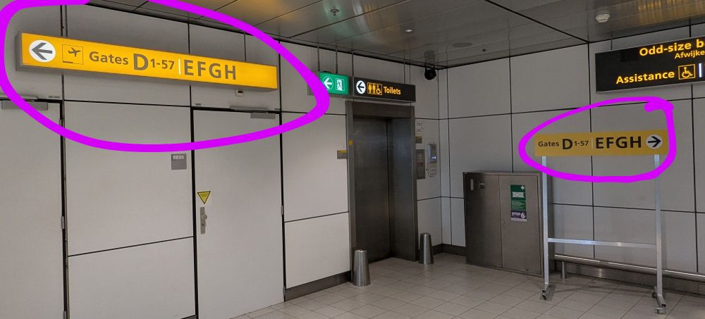 Directions signs at Amsterdam airport pointing in opposite directions for the same destination.