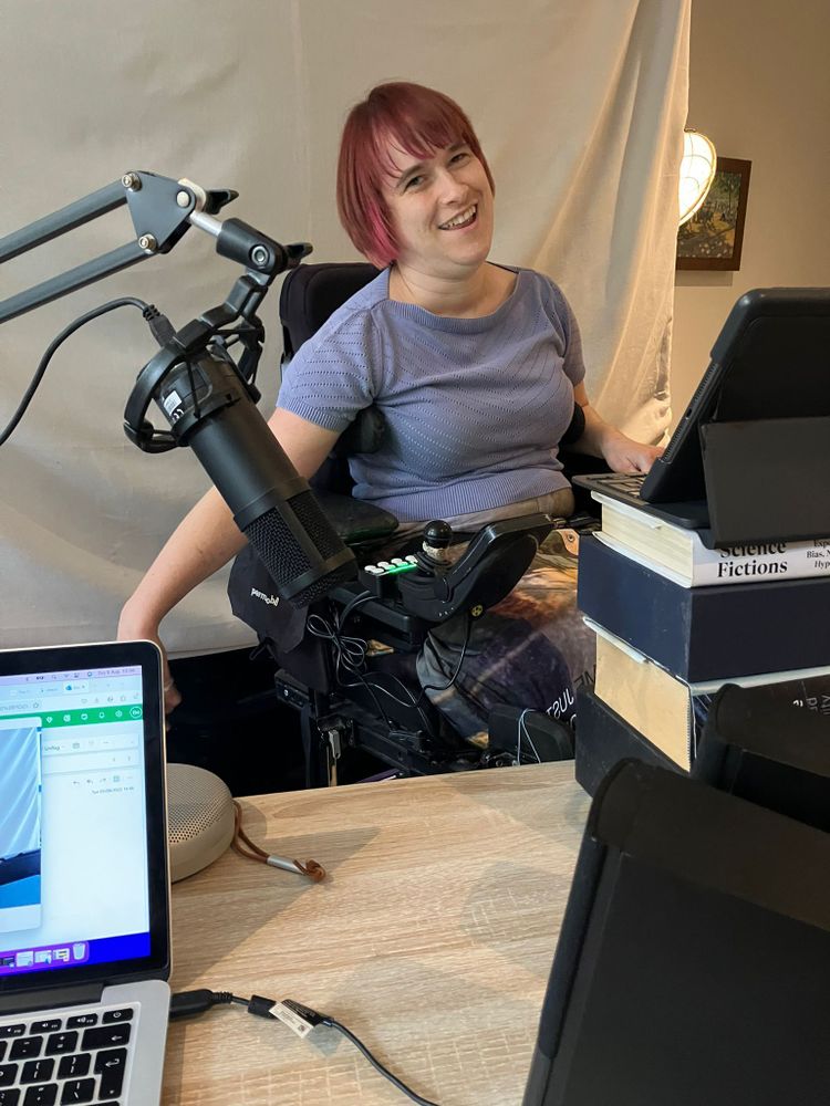 Alt text:
A woman with short red hair smiles while sitting in a powered wheelchair at a home studio desk. She wears a light purple top and is surrounded by tech equipment, including a microphone on an adjustable arm, a laptop, and a tablet propped up on a stack of books. A white sheet hangs behind her as a makeshift backdrop.

