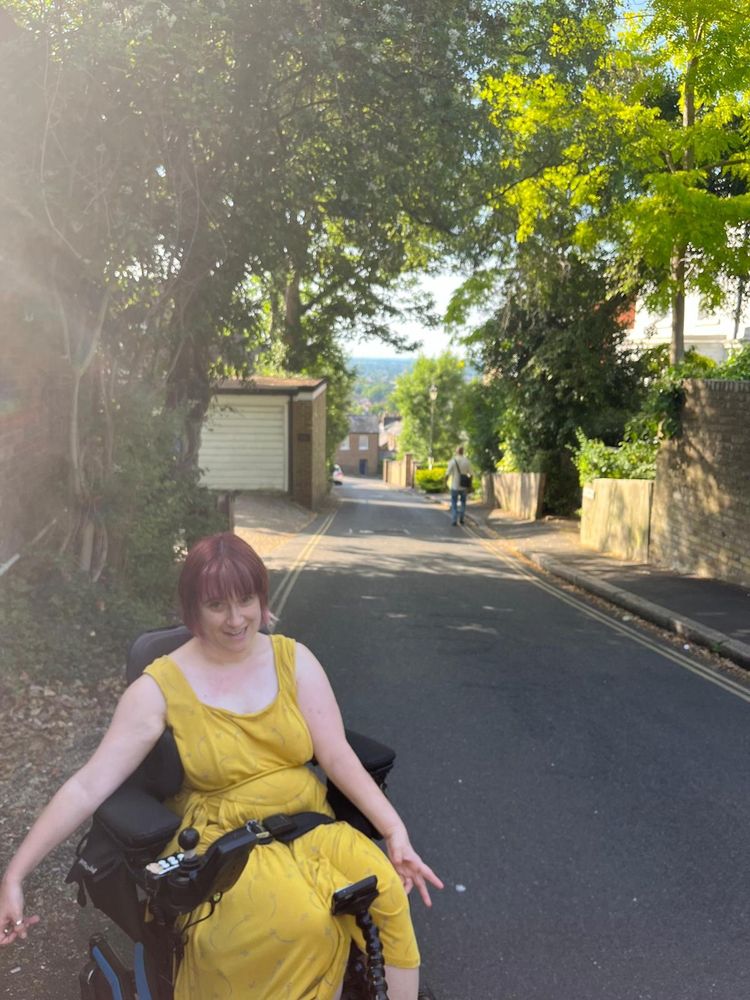 I’m smiling in a yellow dress, sitting in my powered wheelchair on a peaceful, tree-lined street. It’s a sunny day, and you can see someone walking in the distance down the hill, with houses and trees stretching toward the horizon behind them.