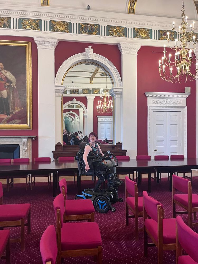 "I am seated in my powered wheelchair in an ornate, historic room with deep red walls, high white columns, and gold detailing. I’m wearing a dark floral dress and smiling toward the camera. The room is furnished with long dark wooden tables and rows of pink-cushioned chairs. A large portrait of a man in regal attire hangs on the left wall, and a grand chandelier hangs overhead. The scene is reflected in a large mirror behind me, adding to the elegance of the setting."








