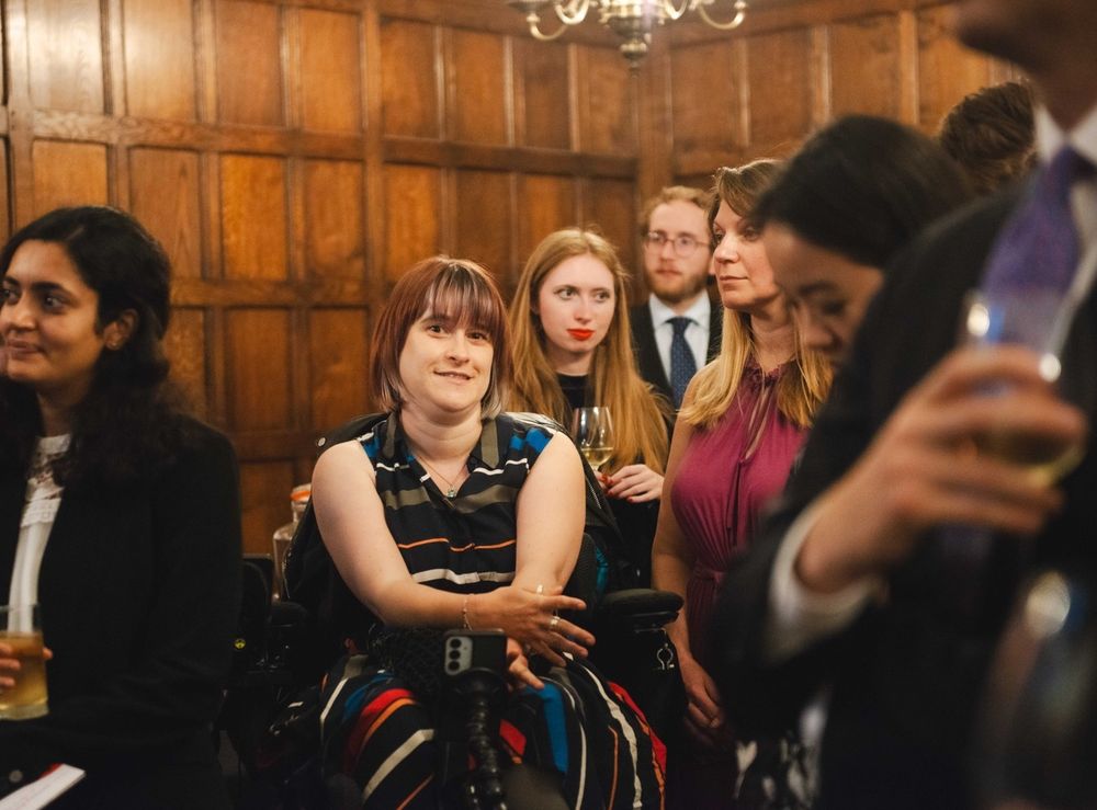 I am at a formal social gathering, seated in my powered wheelchair and smiling. I’m wearing a sleeveless dress with bold, multicolored stripes. Around me, several people in smart attire are engaged in conversation, holding drinks. The room has wooden paneling and a chandelier, giving it a traditional, elegant atmosphere."

