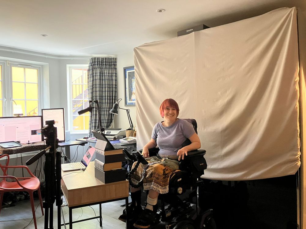 Alt text:
A woman with short red hair sits smiling in a powered wheelchair in a home recording studio. She wears a lilac T-shirt and patterned trousers. Behind her is a large white fabric backdrop, and in front is a desk with multiple monitors, laptops, lamps, and a stack of books used as a camera stand. The room is well-lit with natural light coming through two large windows on the left.

