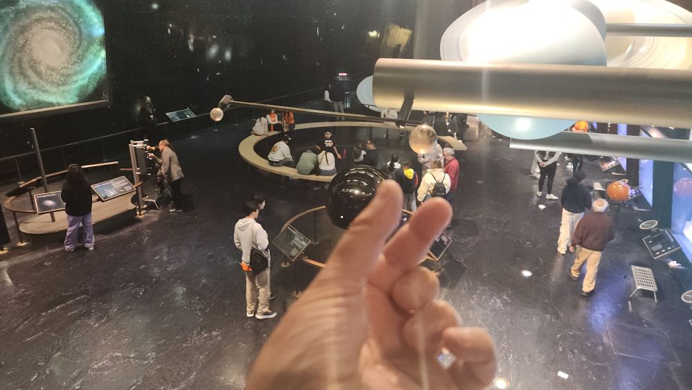 A hand (mine) reaching out to grab a marble sized thing hanging in an exhibit with a bunch of planets in the background. Marbles, obviously, are not planets.