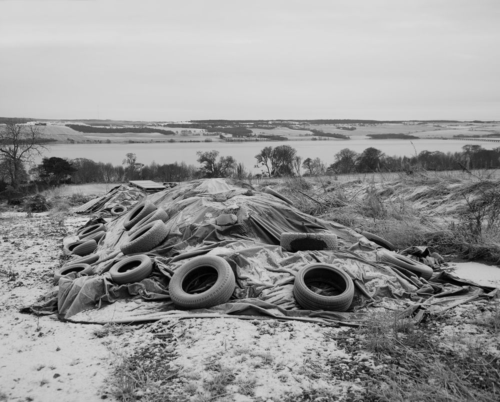 Black & white photograph of a tarpaulin/sheet held down by old tyres, with the Cromarty Firth in the background. Photographed in sharp, frosty conditions.