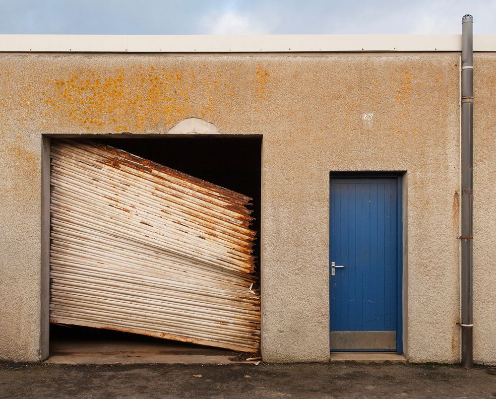 Small industrial unit with a blue door and rusty garage door which is broken and hanging off.