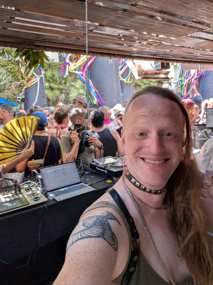 A selfie of a trans woman smiling in a DJ booth, while a large crowd in pride gear dance in the background 