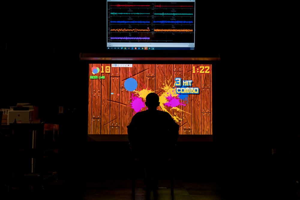 A research participant is seated in a chair in front of a large projector. Projected onto the screen is a game similar to Fruit Ninja wherein the player uses a cursor to slice fruit that appears onscreen. The participant is using their legs to play the game by way of IMU sensors that measure the position of the participant's legs in space and translate that into onscreen cursor movement. The image is taken at the Seáñez biomedical engineering lab at Washington University in St. Louis.