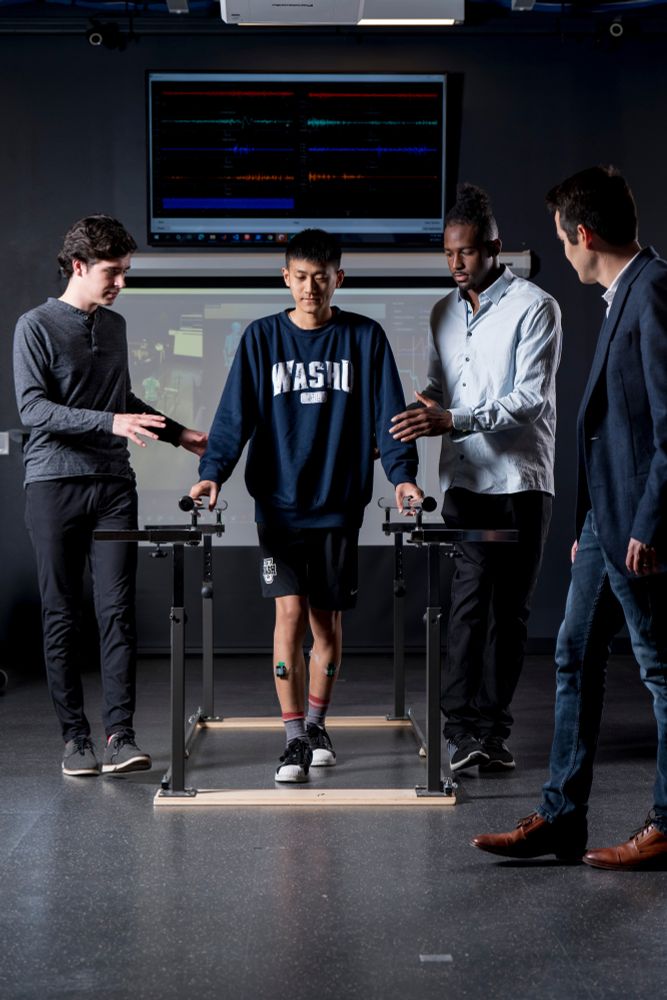 Two researchers help guide a participant during a gait experiment while the principal investigator observes. The participant is wearing sensors on his legs; the image is in the Seáñez biomedical engineering lab at Washington University in St. Louis